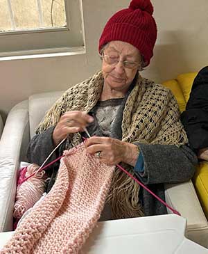 imagem de uma senhora no sofá tricotando uma roupa rosa com uma gorro vermelho na casa de repouso mamma mia