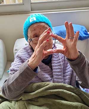 imagem de uma senhora fazendo sinal de coração com as mãos com o gorro azul claro na casa de repouso mamma mia